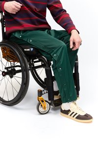 Személy zöld patentos nadrágot, bordó és piros csíkos pulóvert, valamint barna és krémszínű Adidas tornacipőt visel, miközben kerekesszékben ül.