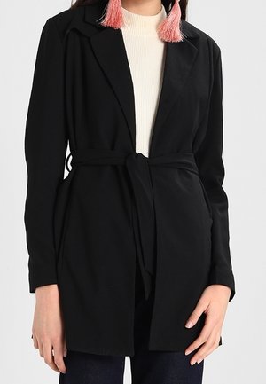 Femme portant un blazer noir ceinturé, un haut blanc à col haut, un pantalon foncé et des boucles d'oreilles à pompons roses, visible du cou aux hanches.