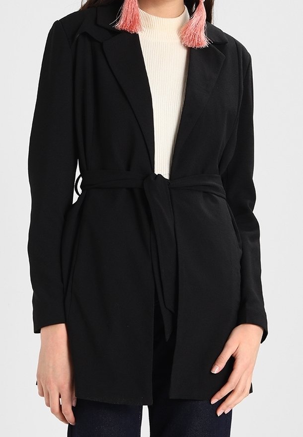 Femme portant un blazer noir ceinturé, un haut blanc à col haut, un pantalon foncé et des boucles d'oreilles à pompons roses, visible du cou aux hanches.