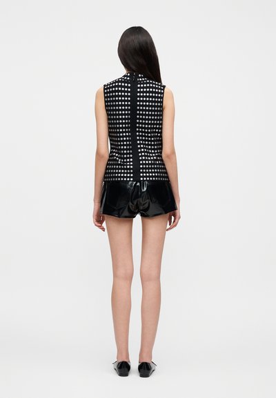 Γυναίκα με μακριά σκούρα μαλλιά, που φοράει μαύρο αμάνικο τοπ με μεταλλικά καρφιά και λαμπερά μαύρα σορτς, στέκεται μπροστά σε απλό φόντο.