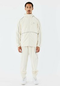 Chándal color crema con chaqueta de cremallera y pantalones ajustados. Presenta detalles en gris, bolsillos con cremallera y puños elásticos. Zapatillas blancas puestas.