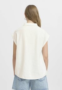Donna con lunghi capelli castano chiaro, che indossa orecchini a cerchio di argento, una maglietta bianca senza maniche e pantaloni corti di denim azzurri, mostrata di spalle.