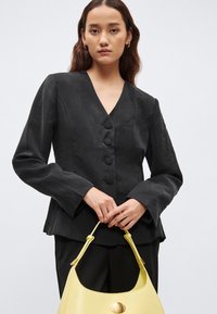 Čierne ľanové sako s výstrihom do V, päť gombíkov na zapínanie a dlhé rukávy. Kombinované so žltou koženou kabelkou s okrúhlym kovovým detailom.