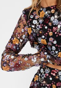 Top in rete nero con maniche lunghe, decorato con ricami floreali e a foglia colorati, con paillettes e dettagli intricati in tutto il capo.