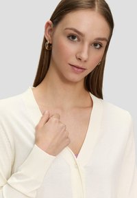 Elfenbeinfarbener Cardigan, gerippte Bündchen und Kragen, weiche Textur, Knopfverschluss, mit einem dezenten Muster, über einem hellrosa Oberteil getragen.