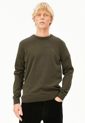 Blonde man die een donkergroene trui met lange mouwen en een zwarte broek draagt, staand met één hand in de zak tegen een lichte achtergrond.