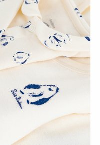 Cremefarbens Kleidungsstück mit Kragen, das blaue Fischmuster und ein gesticktes Logo zeigt. Weiche Textur, klassisches Design.