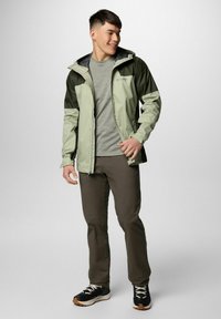 Młody mężczyzna uśmiechający się, ubrany w jasną i ciemnozieloną kurtkę z kapturem, szary T-shirt, brązowe spodnie i czarne trampki z białymi sznurówkami.