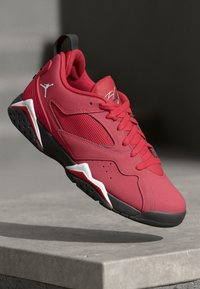 Zapatillas deportivas rojas con una parte superior texturizada, paneles de malla y acentos negros. Presentan un logotipo prominente y un diseño de suela distintivo.