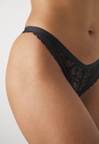 Svart spets-tanga med scallopade kanter och slät midja, som har intrikata blommönster på spetserien.