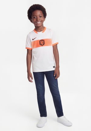 Χαμογελαστό αγόρι που στέκεται, φοράει άσπρη και πορτοκαλί φανέλα ποδοσφαίρου με έμβλημα λιονταριού, σκούρο τζιν και άσπρα αθλητικά παπούτσια σε απλό φόντο.