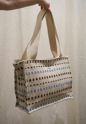 Hand die een beige geweven draagtas vasthoudt met blauwe en bruine gestikte strepen en twee brede stoffen handvatten tegen een lichte achtergrond.