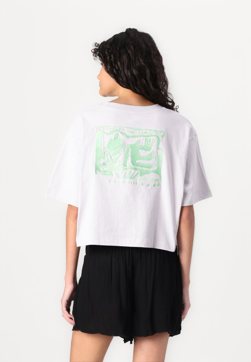Weißes, verkürztes T-Shirt mit einem grünen Grafikdesign auf der Rückseite. Der Stoff wirkt weich und hat eine lockere Passform. Kombiniert mit schwarzen Shorts.