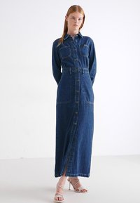 Vestido maxi de mezclilla azul profundo con botones en el frente, mangas largas, dos bolsillos en el pecho, cinturilla y detalle de abertura lateral. Textura suave.