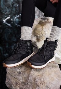 Black high-top sneakers with a padded design, textured fabric, white sole, and decorative laces feature reflective accents.