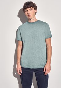 Camiseta de manga corta en un azul claro con cuello redondo; tejido texturizado y sutil detalle del logo en el pecho; combinada con pantalones oscuros.