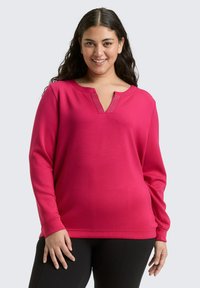 Lächelnde Frau mit langen dunklen Haaren trägt ein leuchtend pinkfarbenes Langarmoberteil mit einem Ausschnitt und schwarze Hosen vor einem hellen Hintergrund.