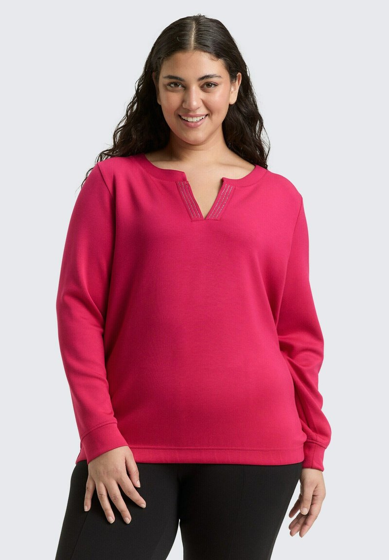 Lächelnde Frau mit langen dunklen Haaren trägt ein leuchtend pinkfarbenes Langarmoberteil mit einem Ausschnitt und schwarze Hosen vor einem hellen Hintergrund.