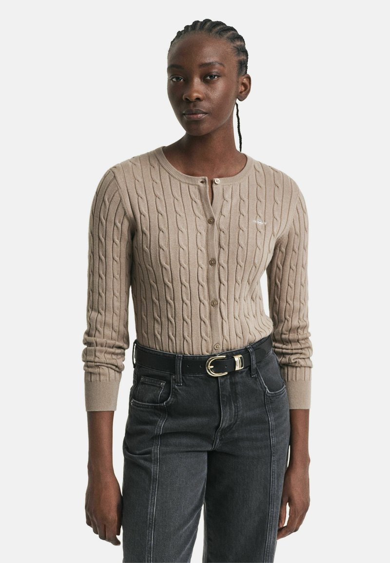 Beige Strickjacke aus Kabelstrick mit Rundhalsausschnitt und Knopfverschluss. Rippbündchen und Saum. Kombiniert mit hochtaillierten schwarzen Jeans und einem schwarzen Gürtel.