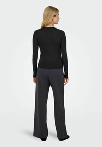 Svart langermet topp med rundet halslinning og bølget kant, kombinert med vide grå bukser. Myk tekstur, tettsittende design.