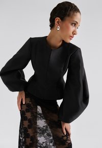 Μαύρο προσαρμοσμένο σακάκι με φουσκωτά μανίκια, συνδυασμένο με ημιδιαφανή μαύρη δαντελένια φούστα με λουλουδάτα σχέδια. Λείο υφασμάτινο υλικό.