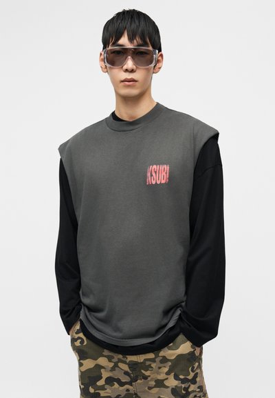 Σκούρο γκρι φούτερ με κοντά μανίκια πάνω από μαύρο πουκάμισο μακρυμάνικο, με κόκκινο λογότυπο. Φορεμένο με σορτς καμουφλάζ και γυαλιά ηλίου.