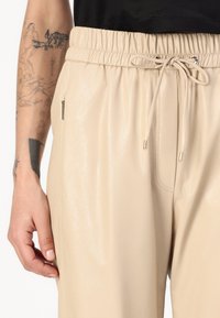 Beige neplederen broek met een elastische tailleband, trekkoord en zijzakken met zilverkleurige ritsaccenten. Gladde textuur.