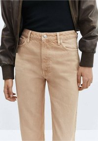 Jean en denim beige à coupe droite, coutures visibles et fermeture classique par bouton. Associé à un haut noir et une veste bomber marron.