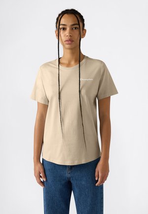 Frau mit langem geflochtenem Haar, die ein beiges Champion-T-Shirt und blaue Jeans trägt, steht vor einem weißen Hintergrund.