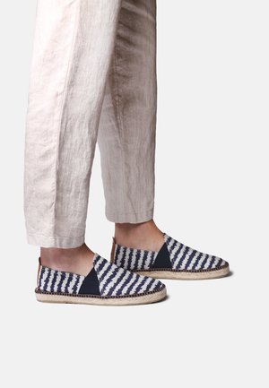 Fødder iført blå og hvide stribede slip-on espadrillos kombineret med lyse beige, korte hørbukser.