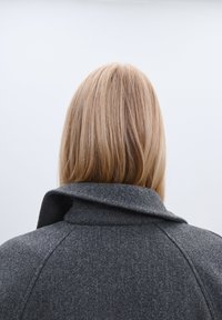 Sivý vlnený kabát s výrazným prehnutým golierom, vyrobený z textúrneho materiálu. Zadný pohľad zobrazuje dlhé, rovné blond vlasy.