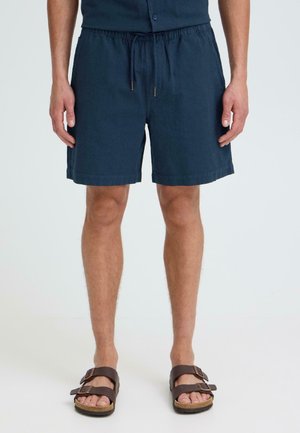 Homme portant un short à cordon de serrage bleu marine et des sandales marron à double sangle, debout devant un fond uni clair.