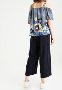 Bluzka z odkrytymi ramionami w kwiatowy wzór w niebieskie, żółte i różowe kolory, z warstwowym designem falbanek, zestawiona z granatowymi spodniami typu wide-leg i białymi trampkami.