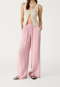 Débardeur beige côtelé avec des boutons, associé à un pantalon large rose clair en tissu texturé et à taille élastique avec cordon de serrage. Sandales noires.