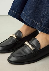 Zwarte lederen instappers hebben een ronde neus en een metallic gouden accent over de bovenkant. De schoenen hebben een vlakke, gestructureerde zool.