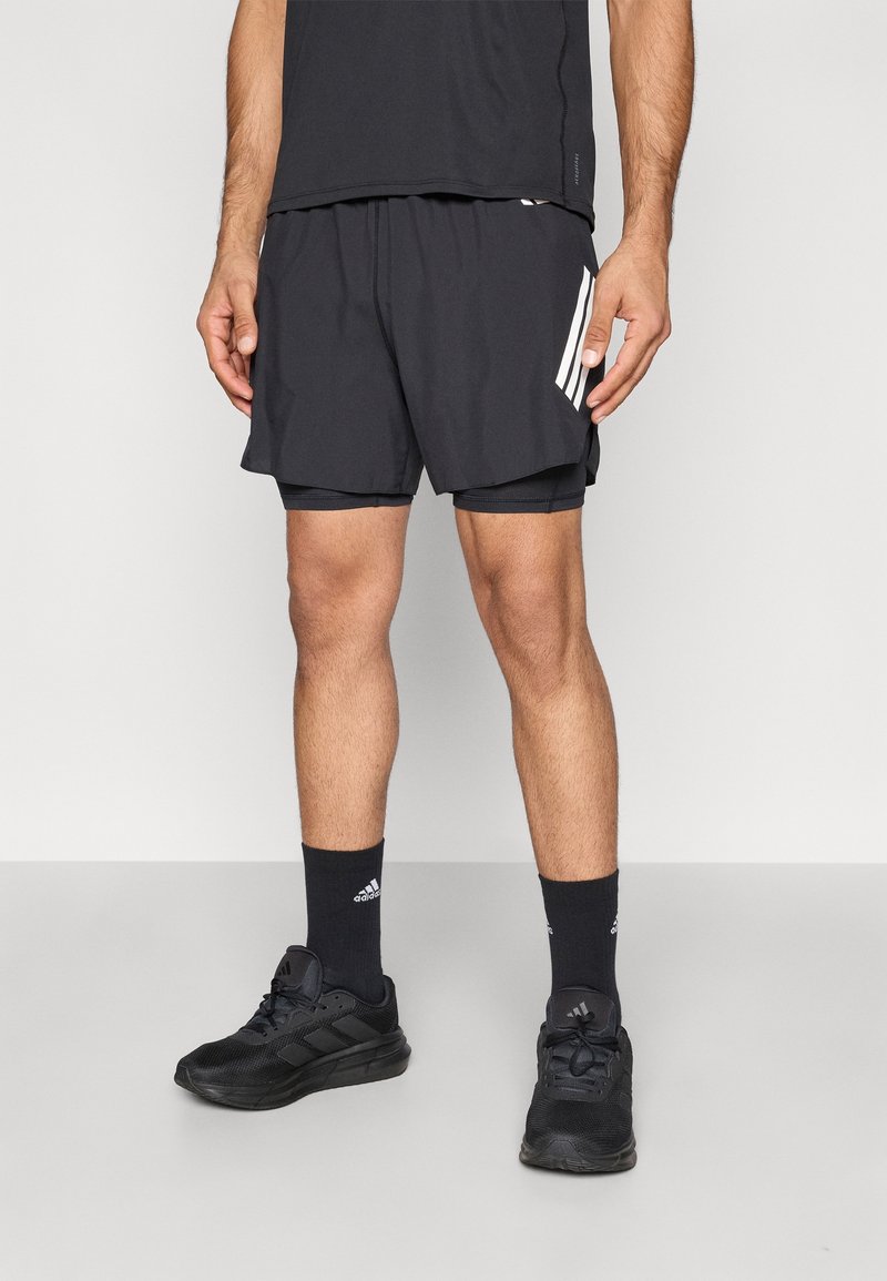 Homme portant un short de sport noir avec des rayures blanches, des chaussures de course en mesh noir et des chaussettes Adidas noires, debout sur un sol gris.
