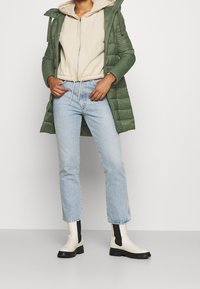 Abrigo acolchado verde sobre una sudadera beige con capucha, jeans rectos de mezclilla azul claro y botas de tobillo off-white con acentos negros y suelas gruesas.