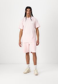 Helles rosa Baumwoll-T-Shirt und passende Shorts, mit lockerer Passform und schlichtem Design. Kombiniert mit hellen Sneakern und weißen Socken.