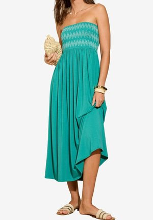 Robe bustier turquoise avec un corsage smocké, une jupe fluide et un ourlet asymétrique. Associée à des sandales plates et une pochette tressée.