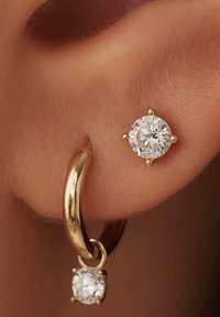 Boucles d'oreilles en or avec un anneau lisse et un composant pendulaire comportant une pierre précieuse claire et ronde, mettant en valeur une surface brillante et des détails complexes.