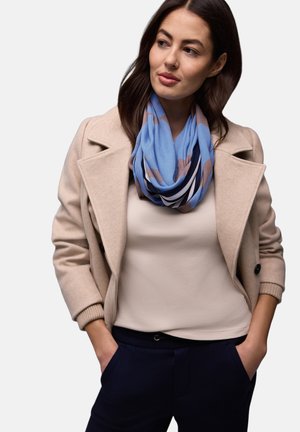 Femme aux cheveux foncés portant un manteau beige, un haut crème, un pantalon bleu marine et une écharpe à motifs bleus et marron, mains dans les poches, regardant sur le côté.