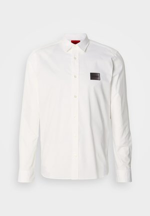 Balta ilgas piedurkņu krekls no kokvilnas, ar apkakli ar pogām, priekšējo uzliktnīti un melnu logotipa plāksteri kreisajā krūtīs.