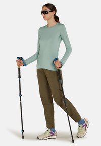 Camicia verde chiaro a maniche lunghe, pantaloni verde oliva e scarpe da trekking multicolore. La persona tiene due bastoni da trekking con dettagli blu.