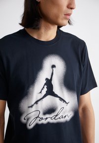 Mustan puuvillapaidan etumuksessa on suuri valkoisella ja harmaalla spraymaalilla piirretty siluetti koripalloilijasta, ja alla on teksti "Jordan". Lyhyet hihat.