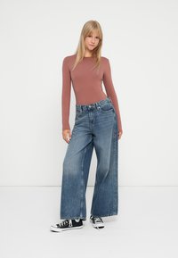 Haut ajusté à manches longues de couleur rouille associé à un jean en denim bleu taille haute à jambes larges et des baskets en toile noires avec des œillets blancs.