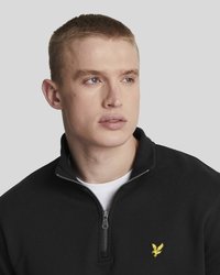 Jongeman met kort blond haar, gekleed in een zwart sweater met een kwart ritssluiting en een klein geel arendlogo, kijkt naar rechts.