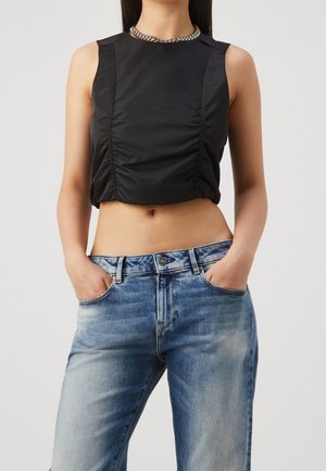 Woman wearing a sleeveless black ruched crop top, silver chain necklace, and faded blue jeans with hands in pockets.