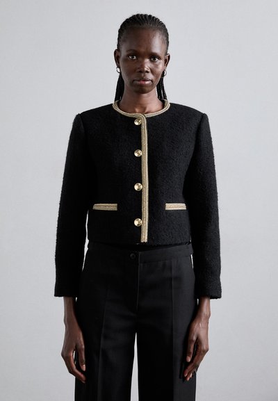Femme portant une veste noire texturée avec des bordures et boutons dorés, associée à un pantalon noir, se tenant devant un fond gris clair uni.