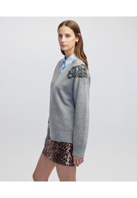 Grijze v-hals trui van gebreid materiaal, met decoratieve versieringen op de schouder. Gecombineerd met een lichtblauw overhemd met kraag en een rok met luipaardprint.