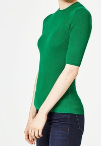 Zelený žebrovaný pletený top s krátkým rukávem a kulatým výstřihem, přiléhavého střihu a hladké textury. Nosený spolu s tmavými džíny.